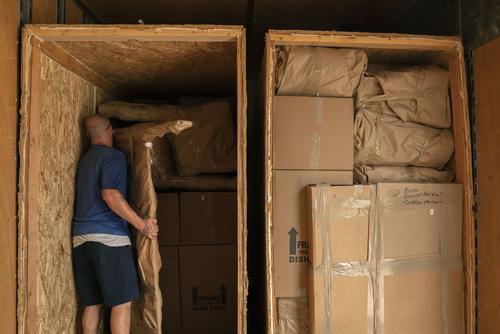 A mover in a T-shirt and shorts fits a brown-paper-wrapped bed frame into a tall crate packed with other brown-wrapped belongings.