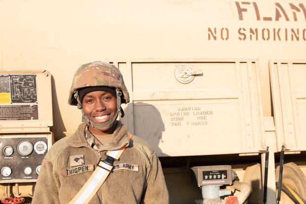 A female soldier stands in front of a truck. 