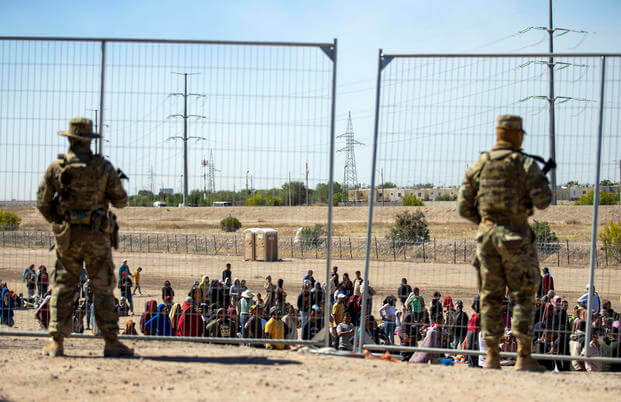 Migrants wait in line adjacent to the border fence under the watch of the Texas National Guard to enter into El Paso