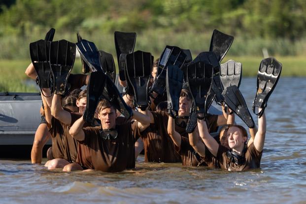 A grouping of students in brown T-shirs wade through chest-high river water holding diving flippers over their heads.