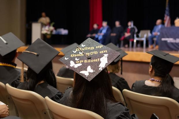 Pictured from an auditorium seat, a graduate's hat decorated with silhouettes of butterflies and a high-heeled graduate, includes the words "Giving up was not an option," during a graduation ceremony with a panel of speakers in the background.