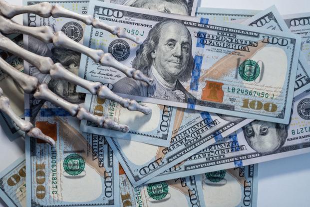 Fingers of a skeleton hand reach into the frame to touch a scattered pile of $100 bills.