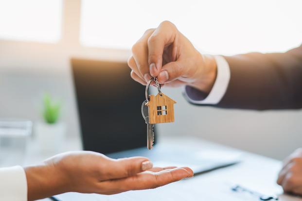 A hand of a man wearing a suit is poised to place a key attached to a house-shaped keychain into a woman's open palm.