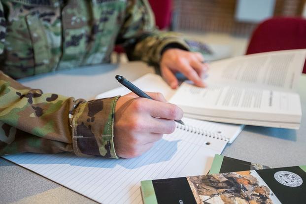 The chest and arms of a soldier in a camouflage uniform, one hand holding a pen, appear poised over a textbook and note paper.