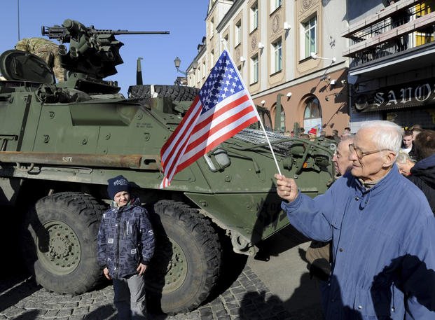 Man holding a US flag walks past a US Army Stryker