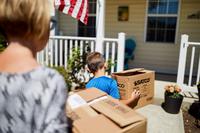 Pictured from behind, a woman and boy each carry a cardboard box toward a yellow house with a white railing and American flag, implying the house is part of military family housing.