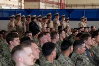 Some uniformed attendees in an aircraft hanger stand and salute while others sit in chairs.