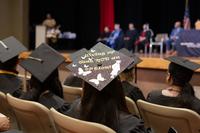 Pictured from an auditorium seat, a graduate's hat decorated with silhouettes of butterflies and a high-heeled graduate, includes the words &quot;Giving up was not an option,&quot; during a graduation ceremony with a panel of speakers in the background.