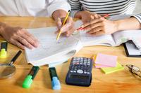 A pair of hands work at some math with a calculator, highlighters, and post-it notes scattered around.