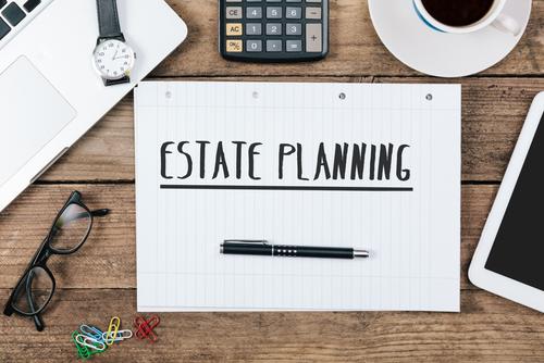 The words &quot;estate planning&quot; appear in black letters sideways on lined notebook papers, on a wood desktop surrounded by office items and coffee.
