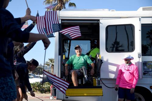 A veteran in a wheelchair exits a van to a crowd waving American flags.